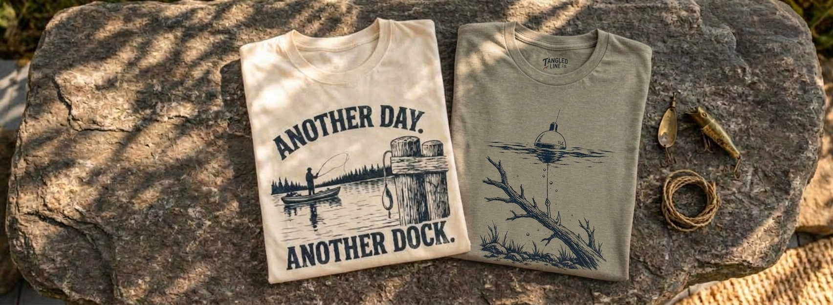 Folded cream and olive fishing graphic tees on a weathered stone surface beside fishing lures and coiled line